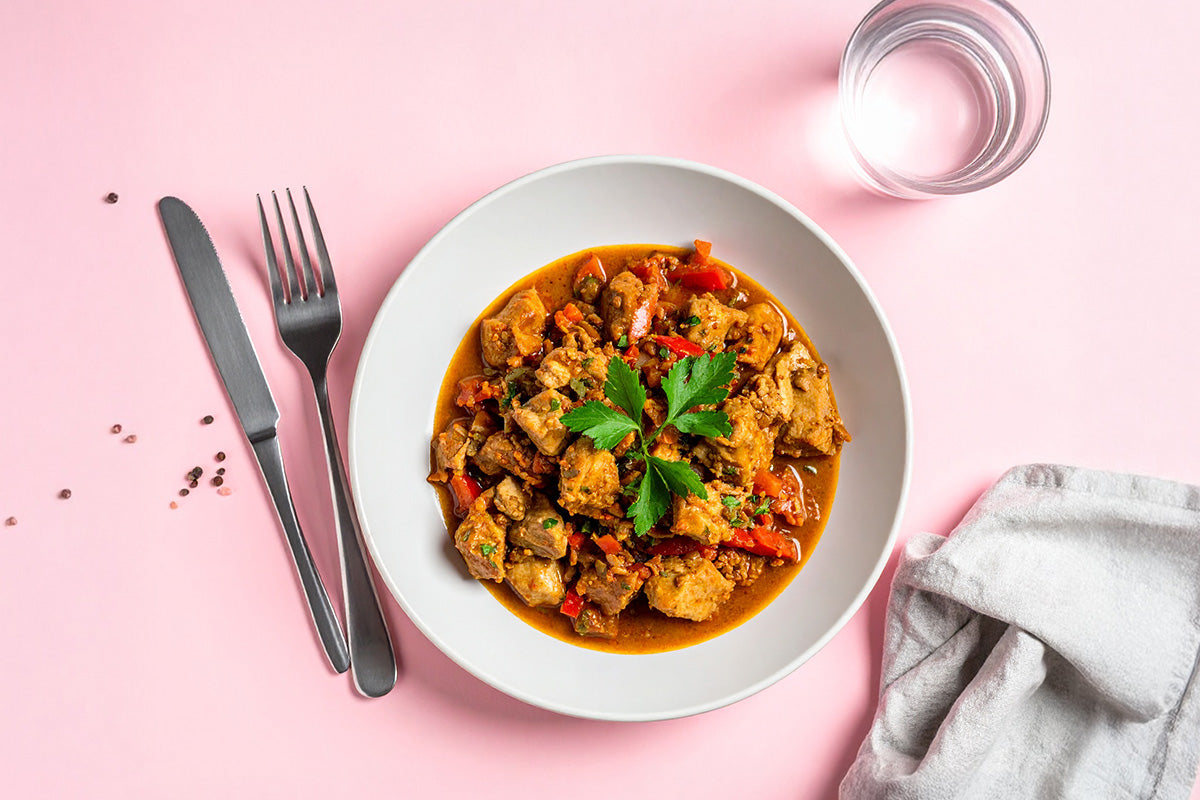 Putenoberkeulengulasch mit Paprika und Petersilie in weißem Teller, daneben Messer, Gabel, Glas Wasser und graues Tuch auf rosa Untergrund