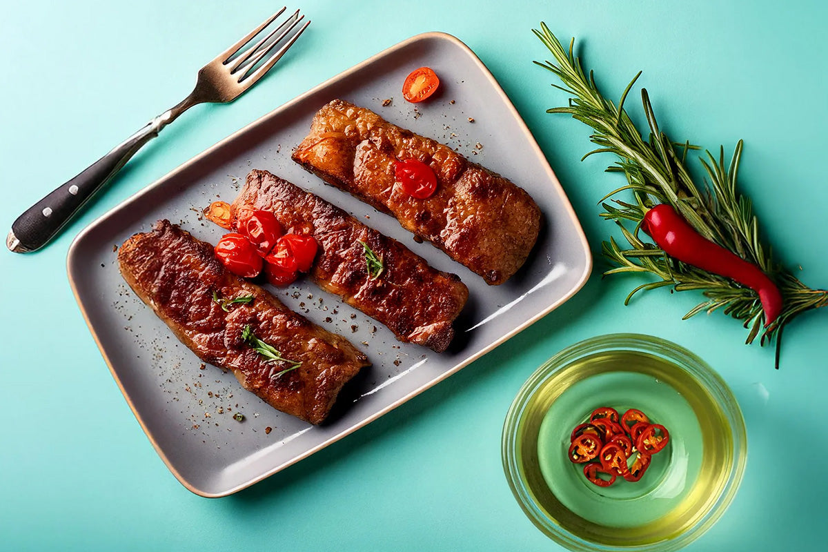 Drei gebratene Hirschsteaks auf einem rechteckigen grauen Teller, garniert mit Kirschtomaten und Rosmarin, daneben eine Gabel, ein Bund Rosmarin mit roter Chili und eine Schale mit Öl und Chilischeiben