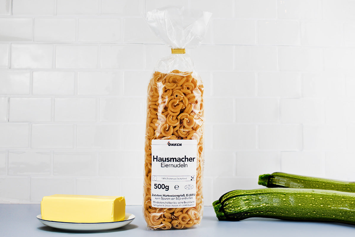 Transparente Verpackung mit braunen Eiernudeln der Marke Hausmacher auf weißem Untergrund neben Butter auf weißem Teller und zwei Zucchini