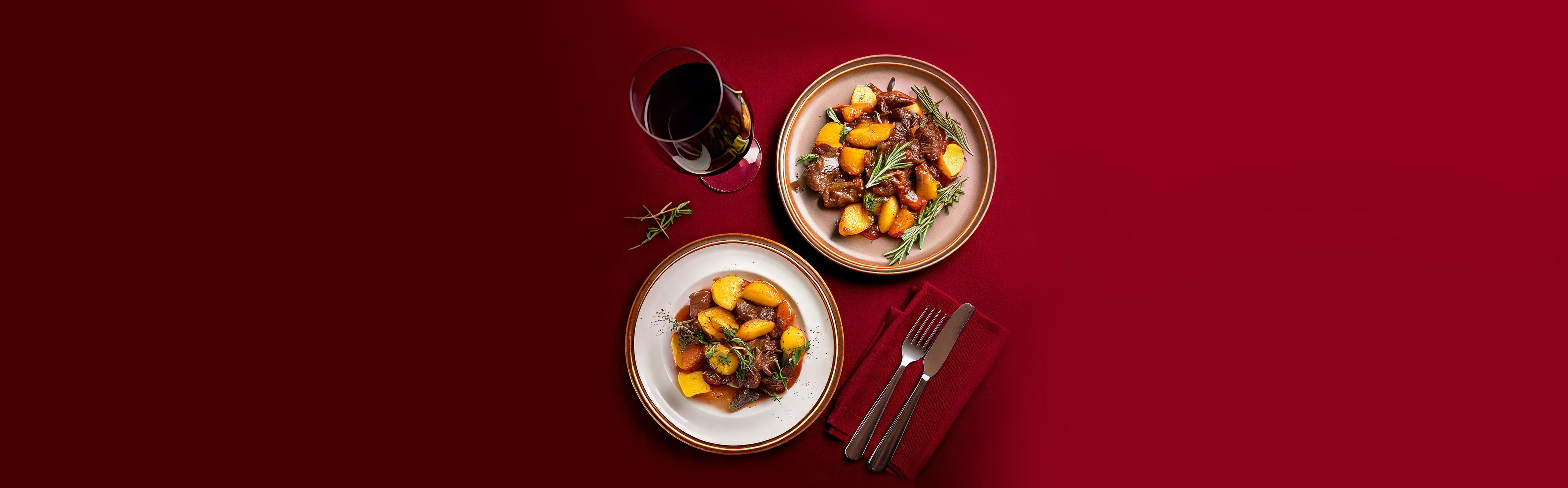 Zwei Teller mit Hirschgulasch, Kartoffeln und Rosmarin, daneben ein Glas Rotwein und Besteck auf rotem Hintergrund