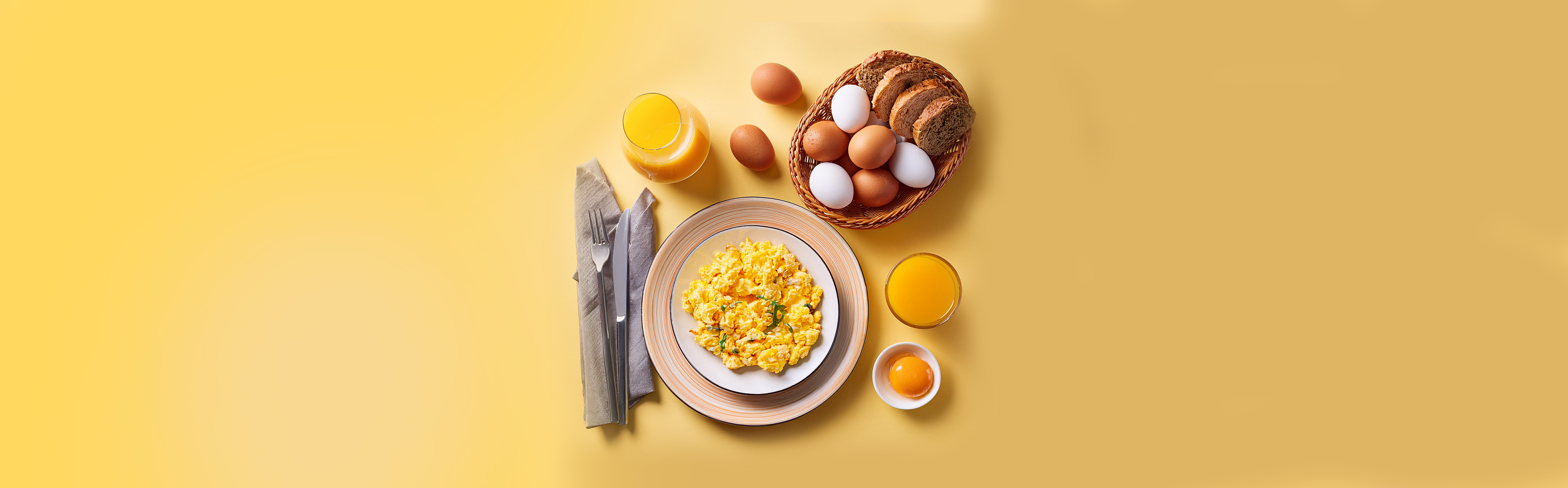Rührei auf weißem Teller mit Besteck, Orangensaft, Brotkorb mit Eiern und aufgeschlagenem Ei auf gelbem Hintergrund