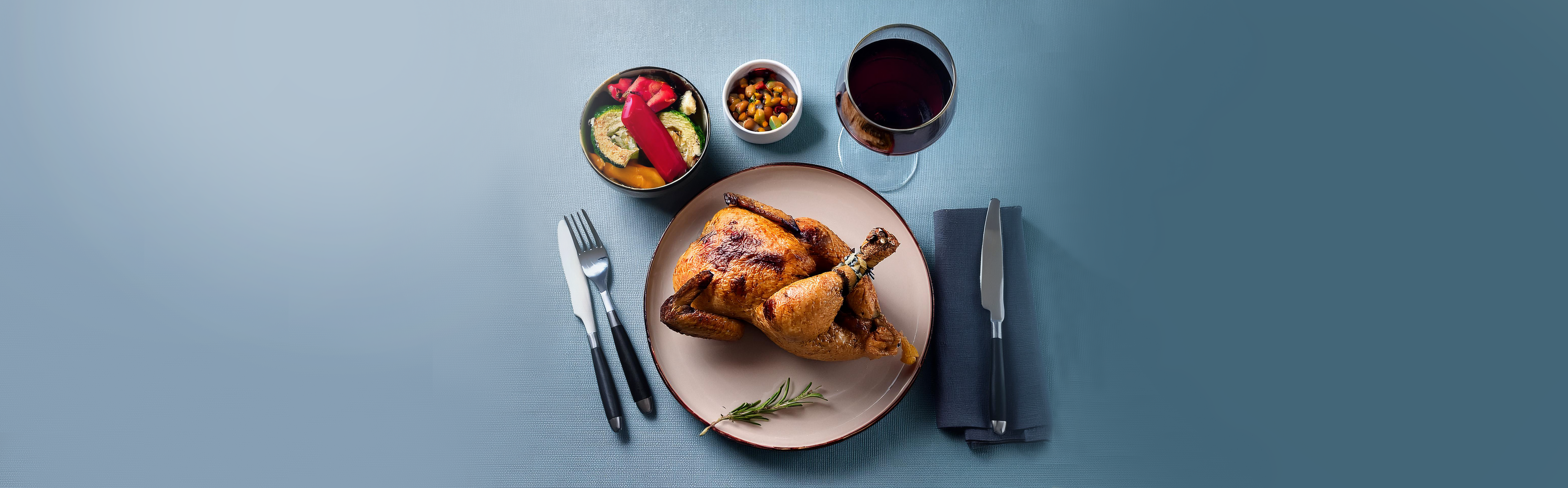 Gebratenes ganzes Huhn auf Teller mit Rosmarinzweig, dazu kleine Schalen mit Gemüse und ein Glas Rotwein, Besteck auf grauem Tisch