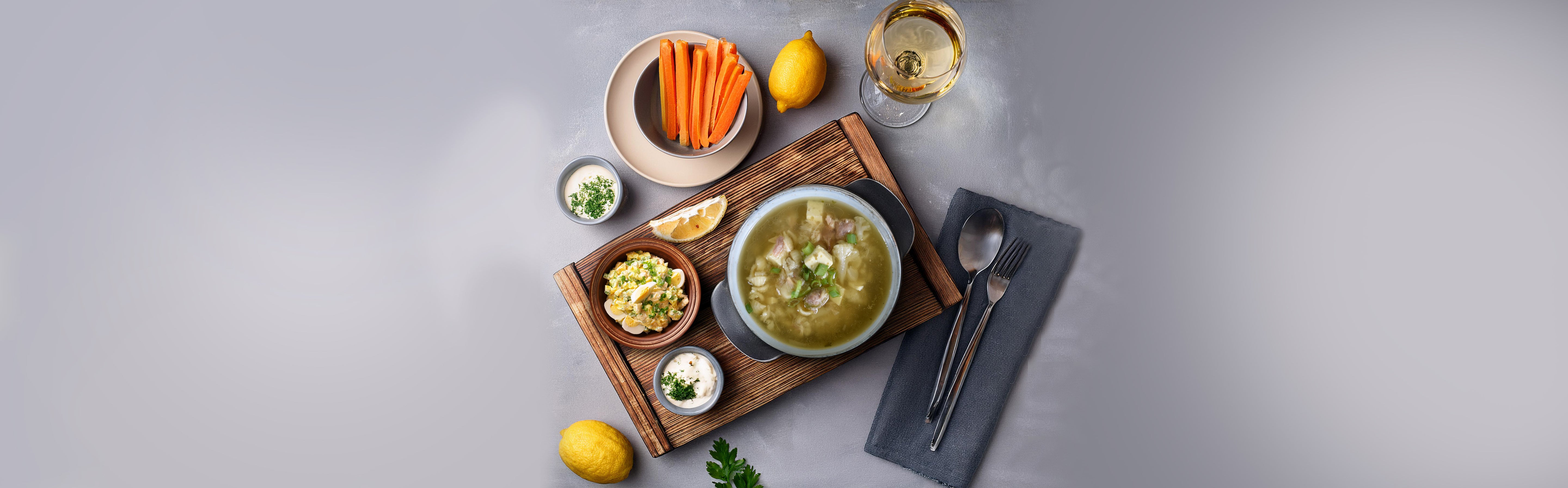 Teller mit Hühnersuppe, dazu Karottensticks, Zitronen, Kräuterdip und ein Glas Weißwein auf grauem Untergrund