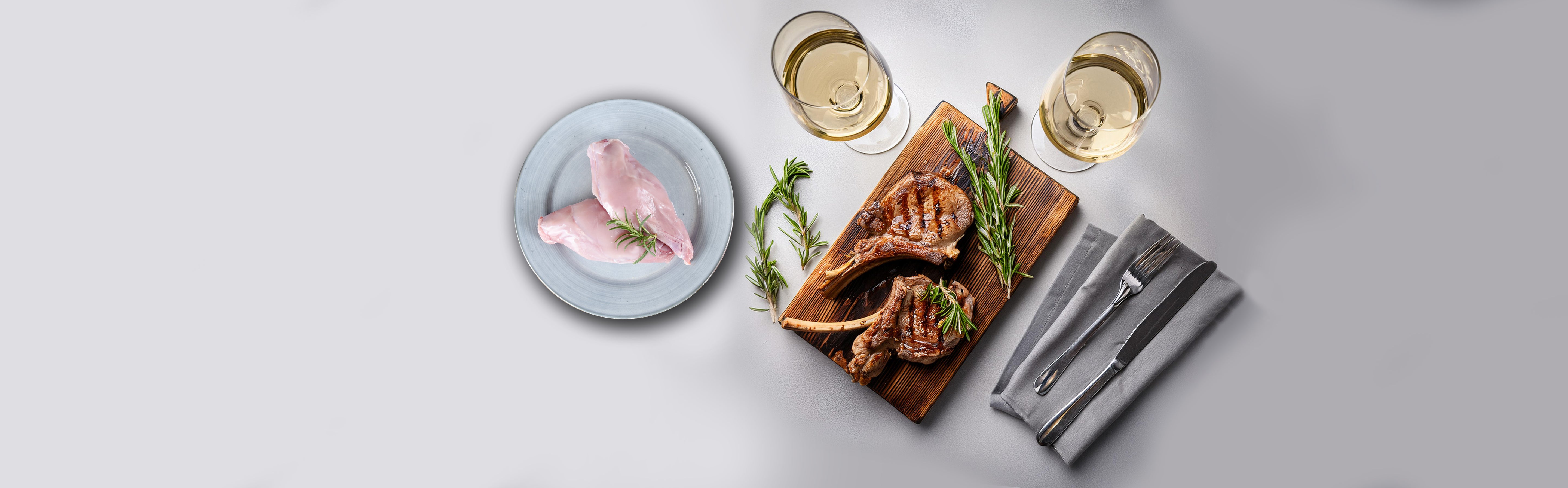 Zwei gegrillte Lammkoteletts mit Rosmarin auf einem Holzbrett, rohes Hähnchenfleisch auf einem Teller, zwei Gläser Weißwein, Besteck auf grauem Stoffserviette