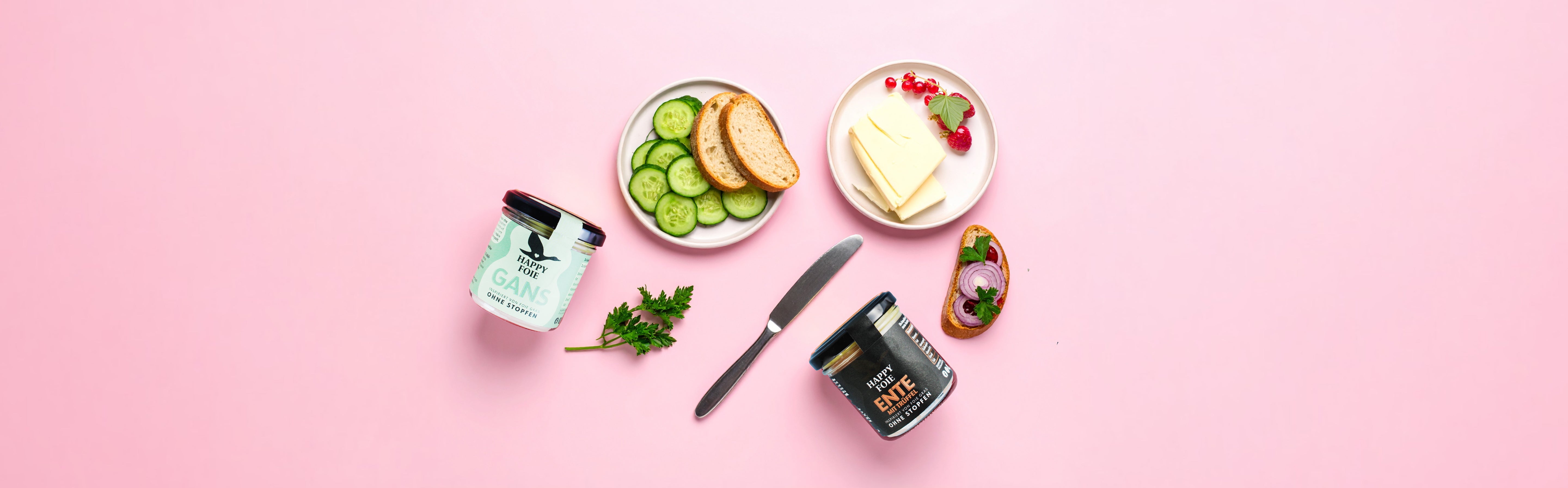 Zwei Gläser Happy Foie Pastete, eine mit Gans und eine mit Ente, daneben Scheiben Brot, Gurken, Butter mit roten Beeren und ein bestrichenes Brot auf rosa Hintergrund