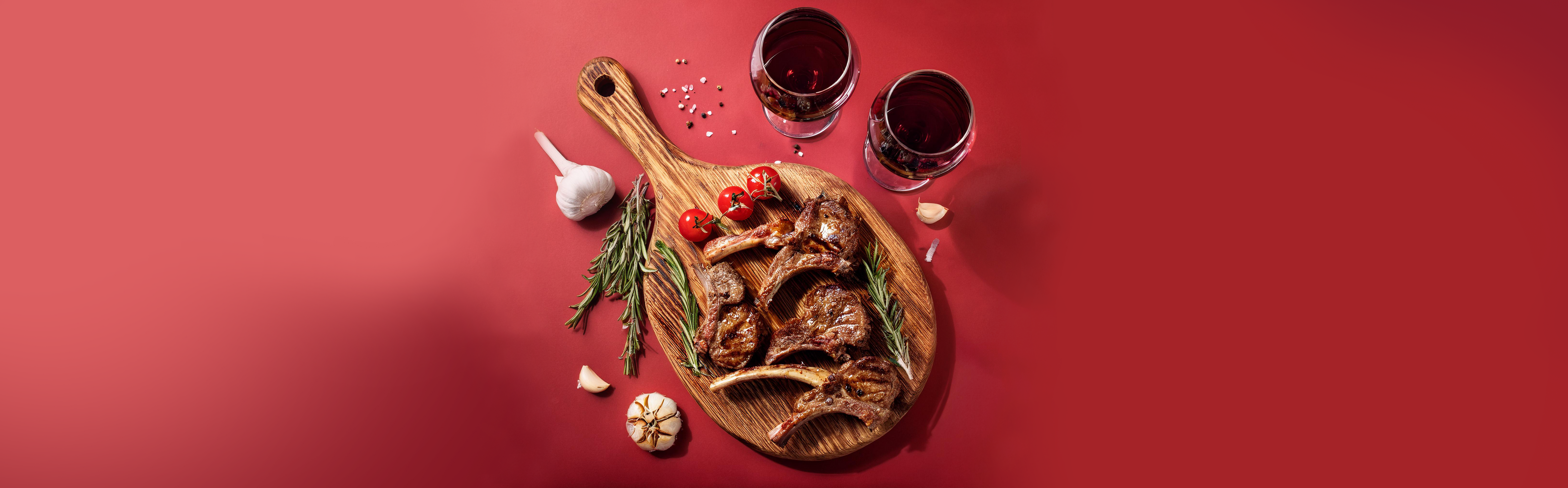 Gegrillte Lammkoteletts auf Holzbrett mit Rosmarin, Knoblauch, Kirschtomaten und zwei Gläsern Rotwein auf rotem Hintergrund