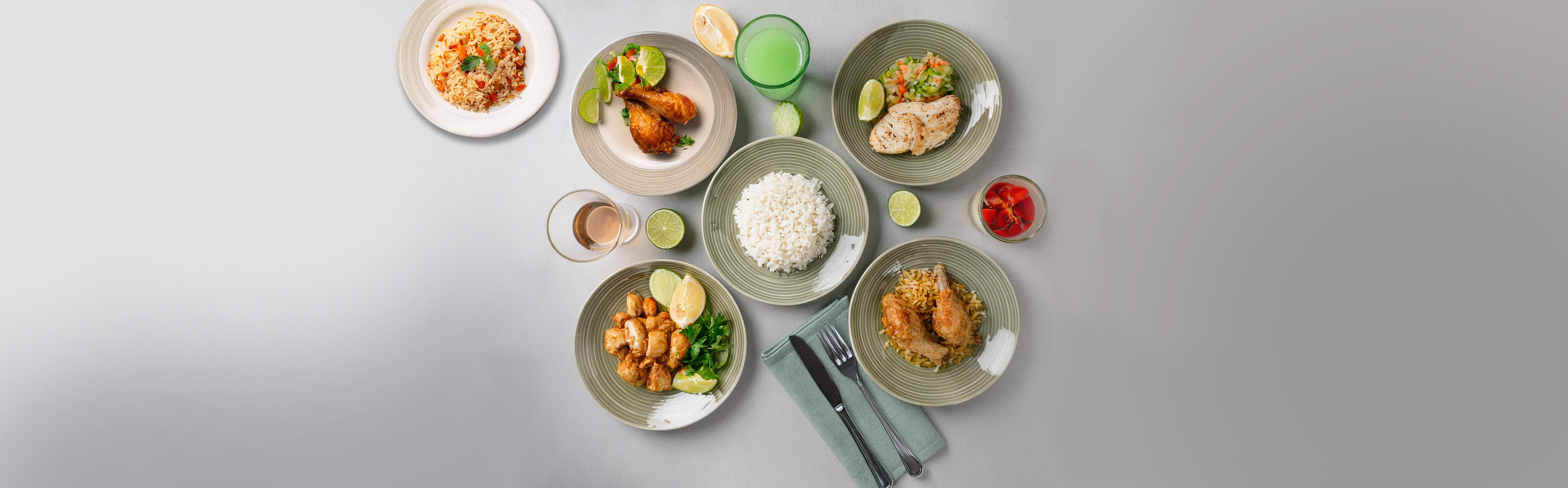 Verschiedene Hähnchengerichte auf Tellern mit Reis, Limettenscheiben und Kräutern, Besteck und zwei Gläser mit Getränken auf grauem Hintergrund