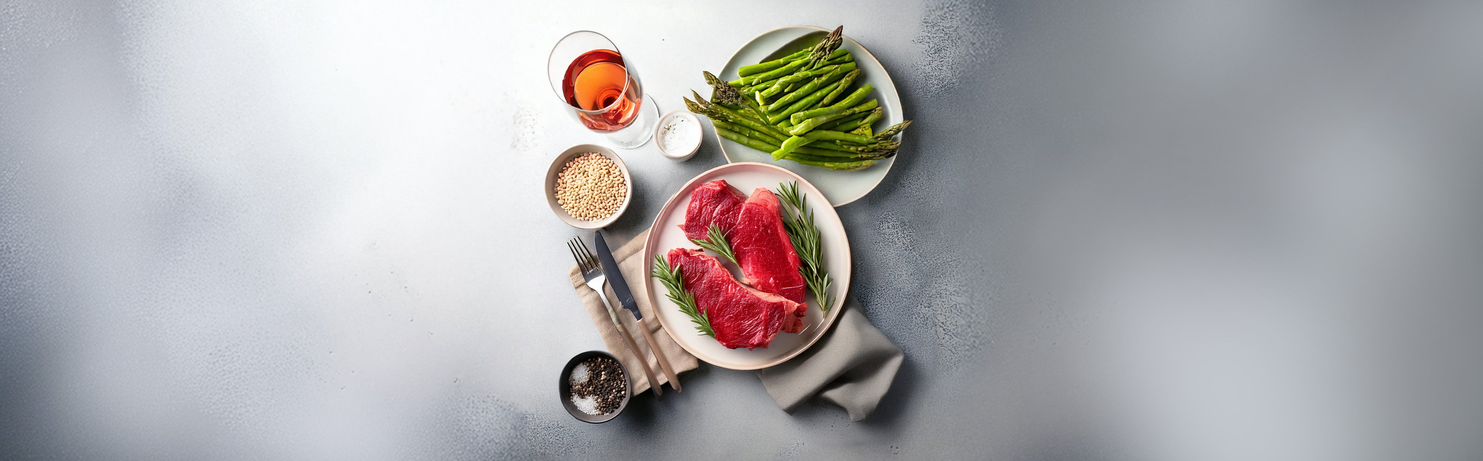 Drei rohe Straußenfleischsteaks auf weißem Teller mit Rosmarin, daneben grüner Spargel auf einem weiteren Teller, Salz, Pfeffer, weiße Bohnen und ein Glas Roséwein auf grauem Untergrund