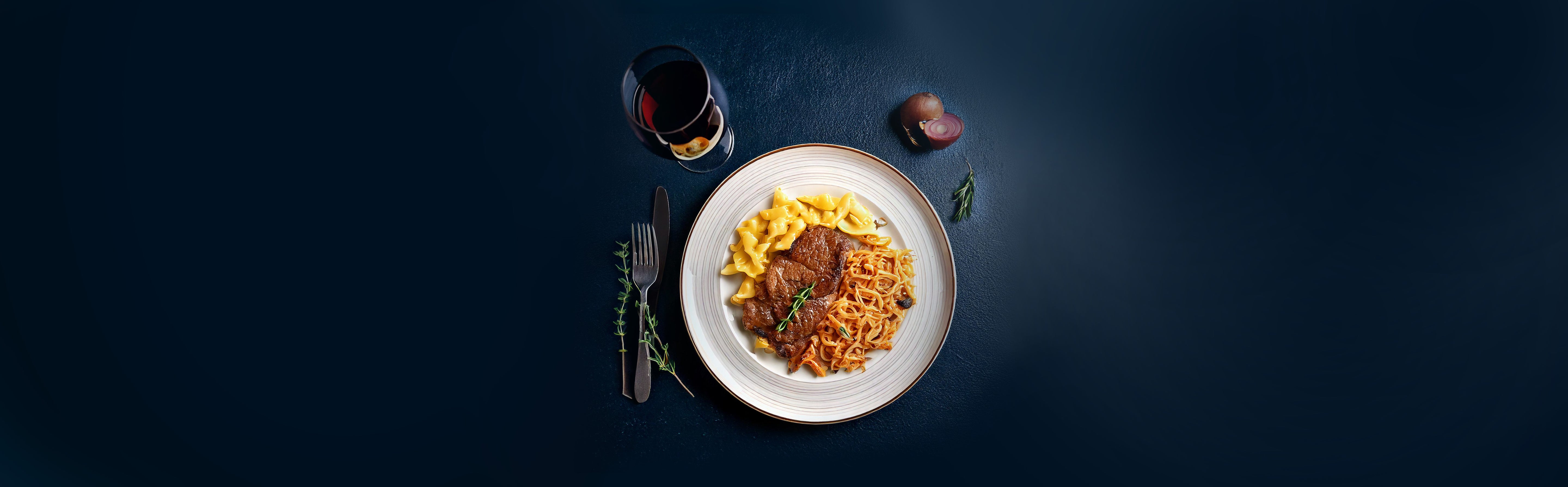 Teller mit Wildschweinbraten, Spätzle und Rotkohl, daneben Messer, Gabel, Glas Rotwein und Zwiebel auf dunklem Hintergrund