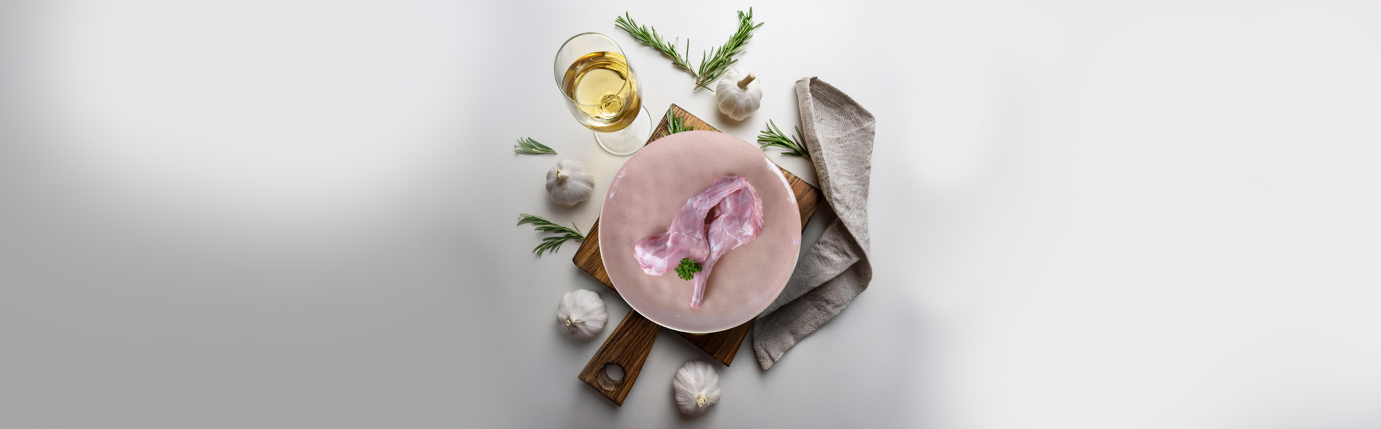Rohe Kaninchenvorderläufe auf rosa Teller mit Knoblauch, Rosmarin, Glas Weißwein und grauem Geschirrtuch auf Holzbrett