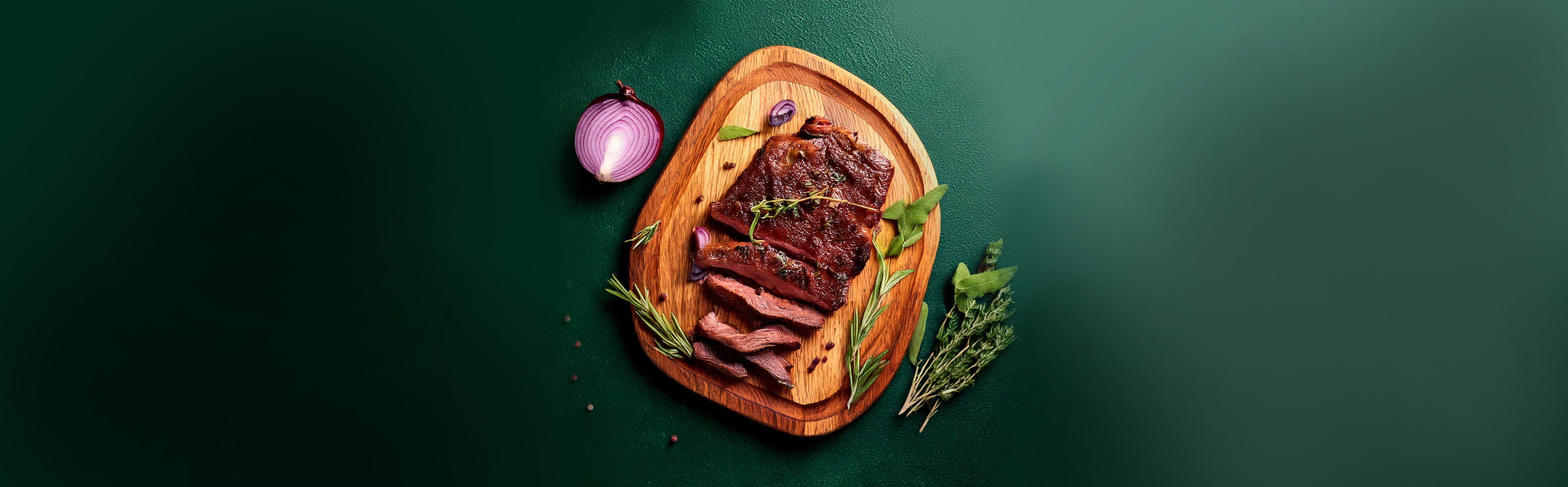 Geschnittenes, gebratenes Rehsteak auf Holzbrett mit Kräutern und halbierter roter Zwiebel auf grünem Hintergrund