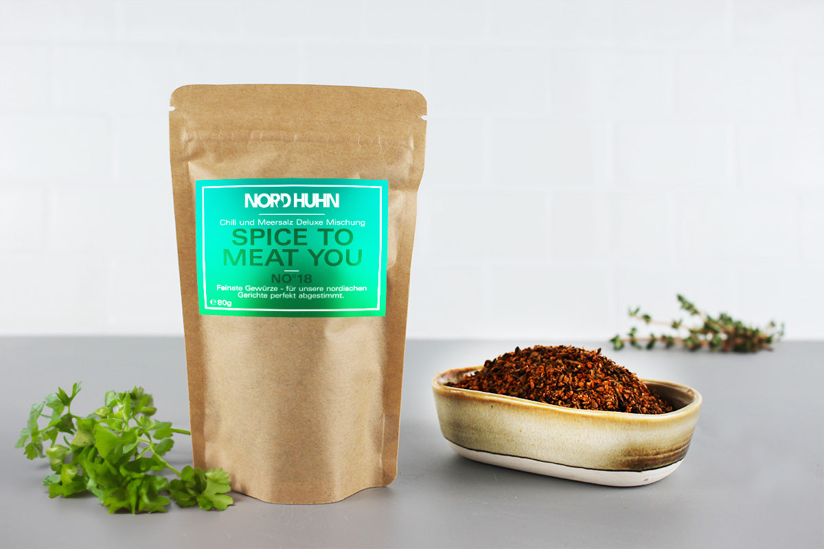 Braunes Gewürzbeutel mit grünem Etikett 'NORDHUHN SPICE TO MEAT YOU' neben ovaler Schale mit braunem Gewürz, grüne Kräuter im Hintergrund.
