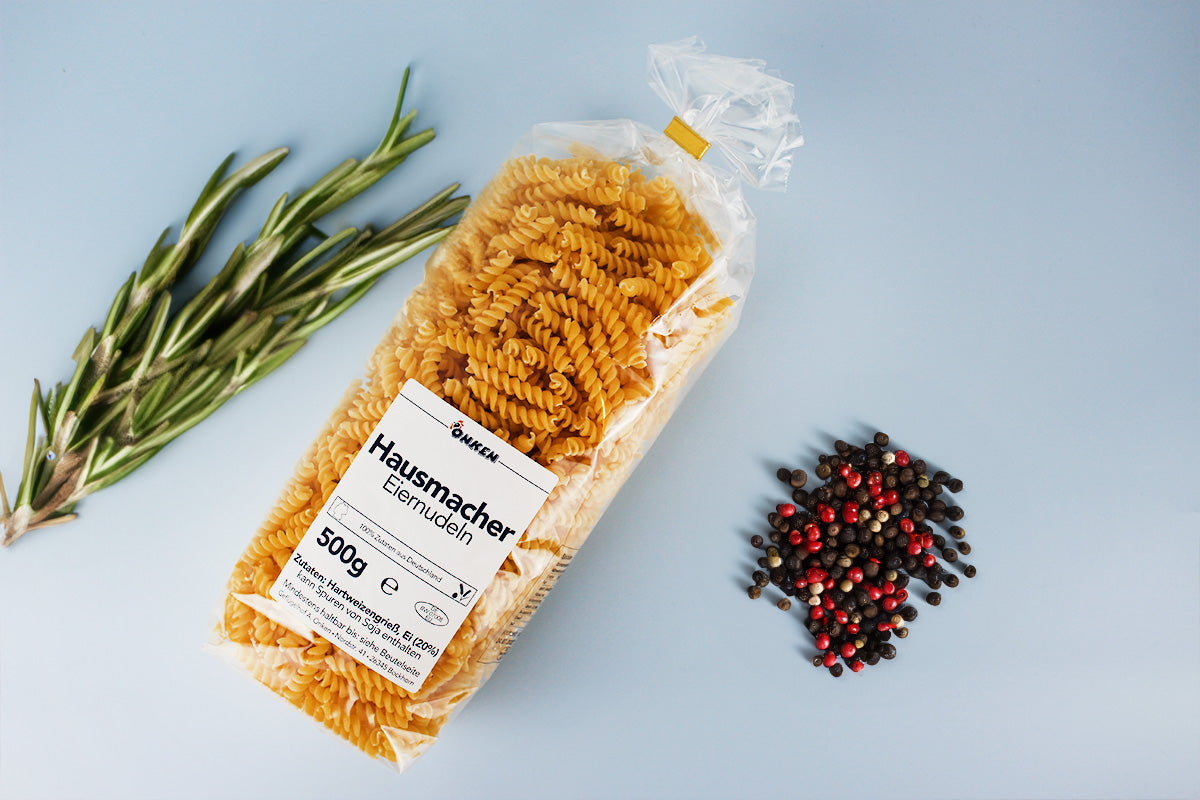 Packung mit Spiralnudeln aus Eierteig neben Rosmarinzweigen und gemischten Pfefferkörnern auf hellem Untergrund