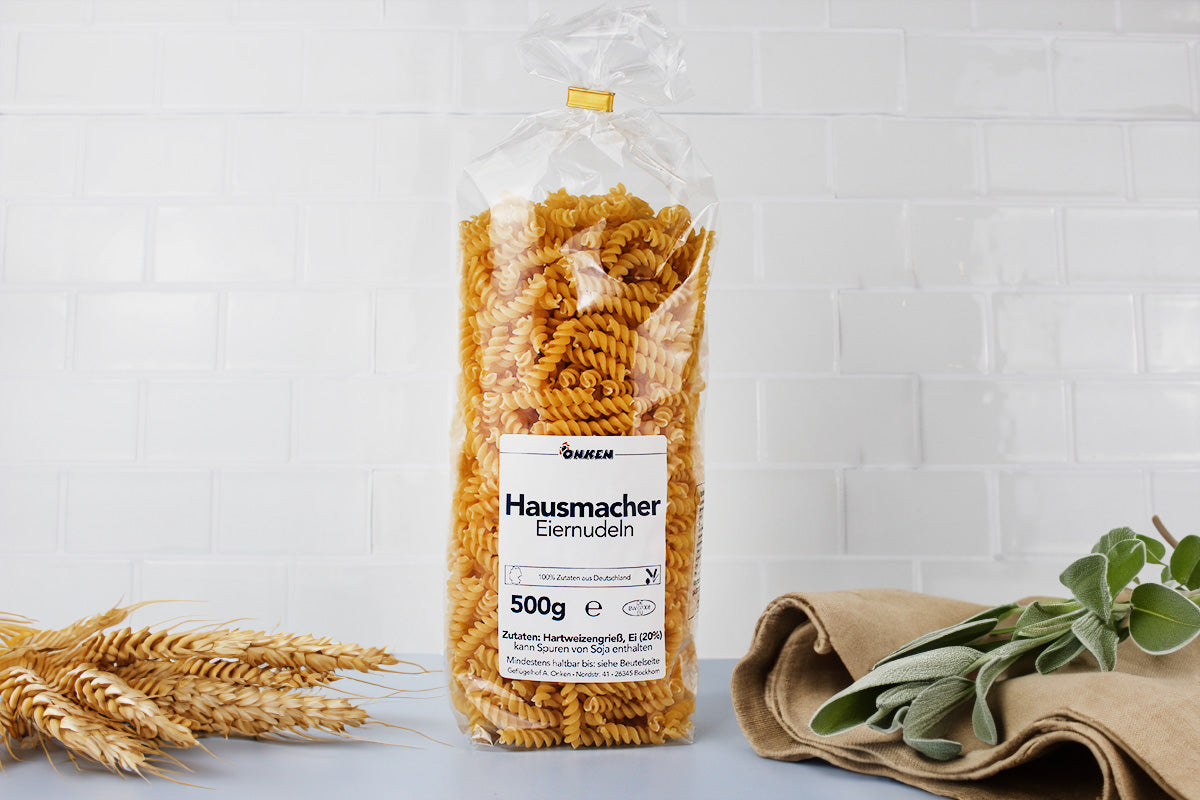 Transparente Verpackung mit Spiralnudeln und weißem Etikett mit der Aufschrift 'Hausmacher Eiernudeln 500g' auf weißem Hintergrund mit Weizenähren und grünen Blättern