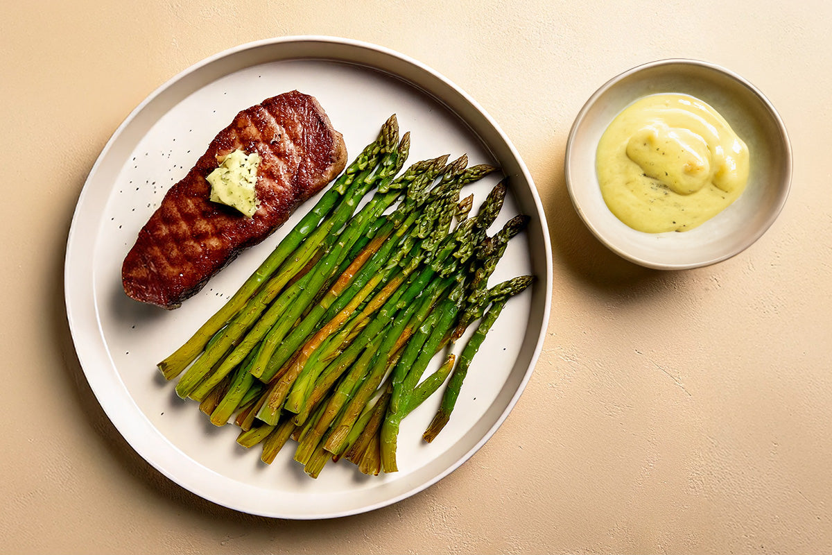Gebratenes Straußenfilet mit Kräuterbutter auf weißem Teller, dazu gegrillter grüner Spargel und eine Schale mit heller Sauce