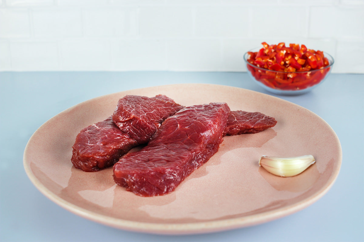 Drei rohe Straußenfleisch-Medaillons auf einem hellen Teller, daneben eine Knoblauchzehe, im Hintergrund eine Schüssel mit gehackten roten Chilischoten