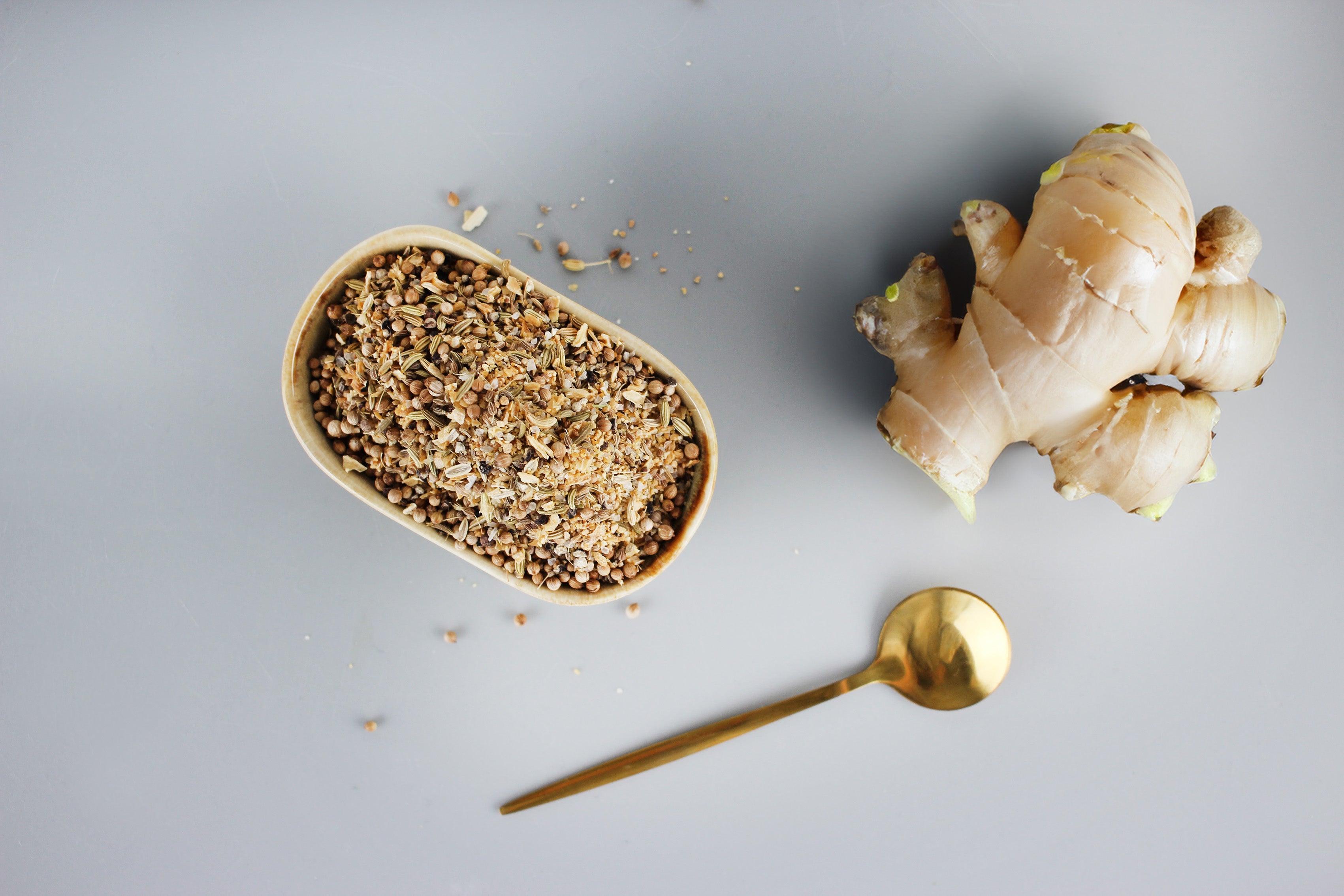 Schale mit gemischten Gewürzen, frischer Ingwer und goldfarbener Löffel auf grauem Untergrund