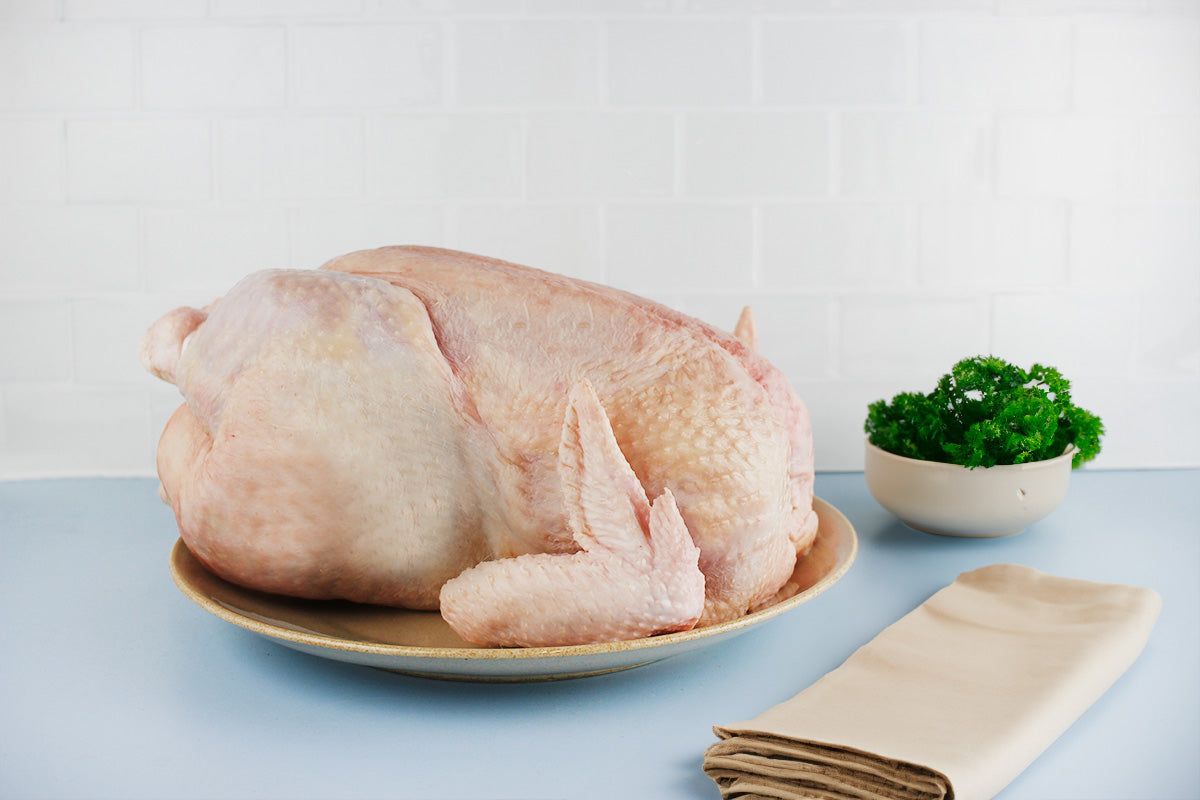 Ganzes rohes Suppenhuhn auf einem Teller, daneben eine Schale mit frischer Petersilie und ein gefaltetes helles Tuch