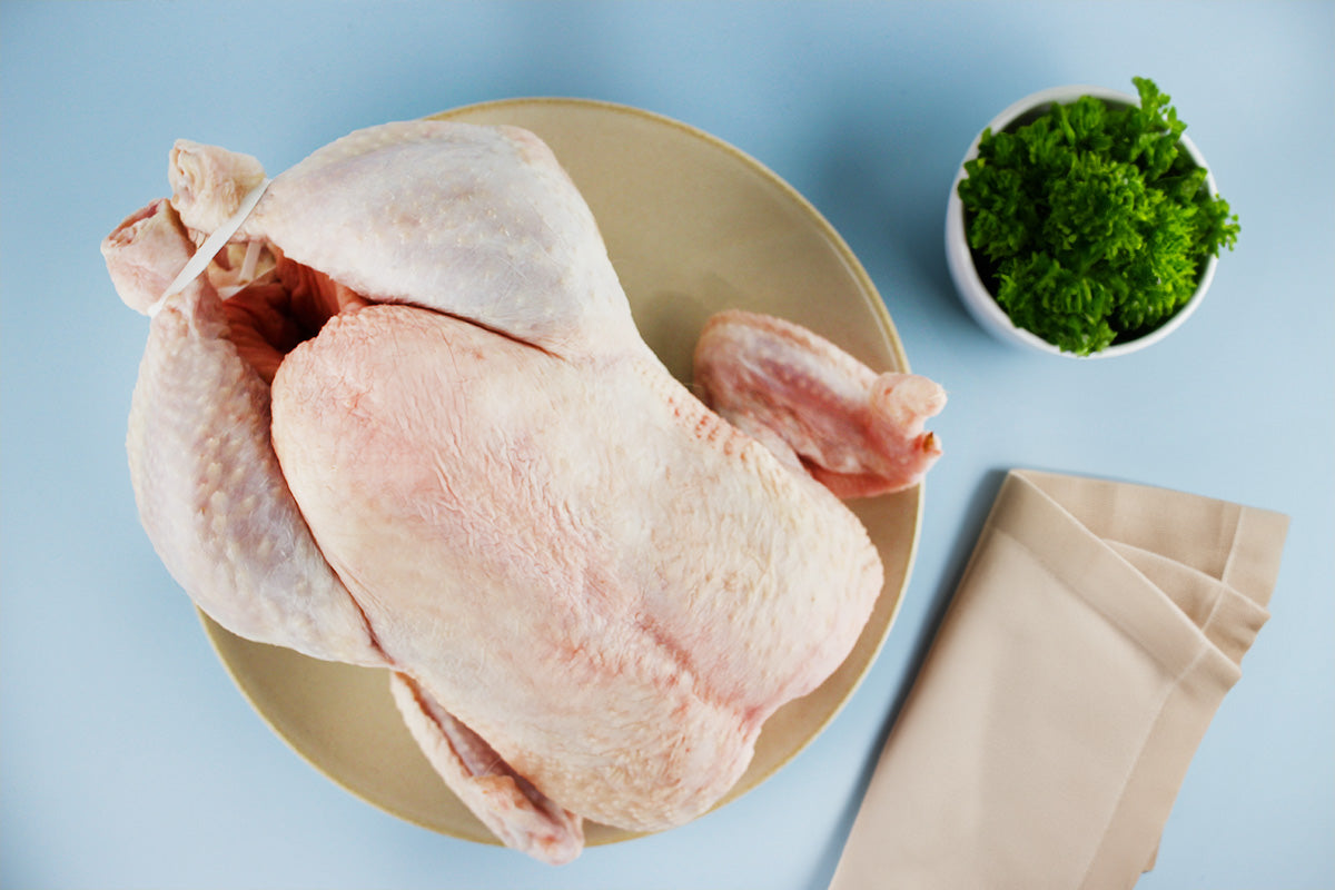 Ganzes rohes Suppenhuhn auf einem Teller, daneben ein Becher mit frischer Petersilie und ein gefaltetes hellbeiges Tuch