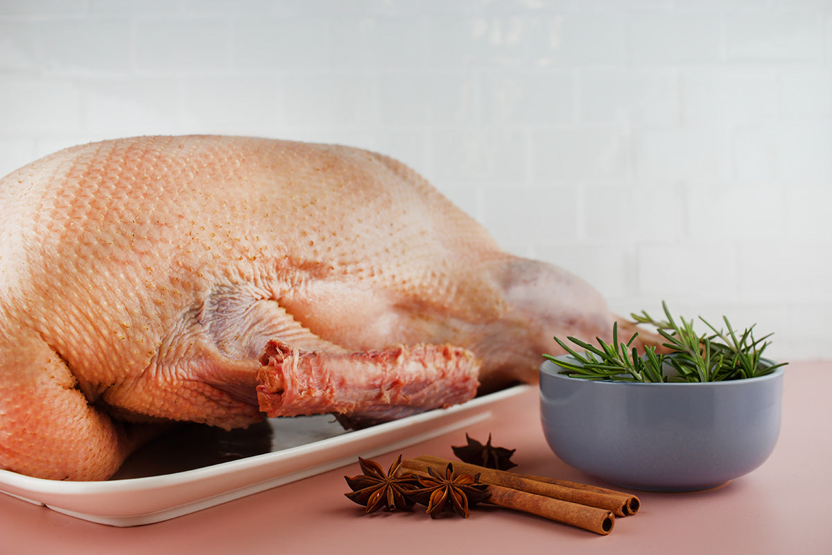 Ungekochte Gans auf weißem Teller, daneben Schale mit Rosmarin und Gewürze wie Zimtstangen und Sternanis auf rosa Untergrund.
