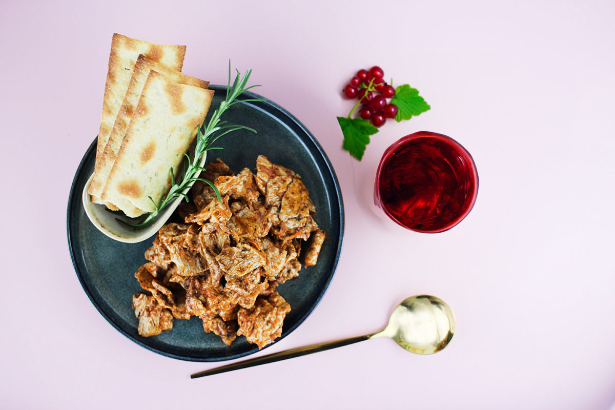 Veganes Gyros aus Soja-Protein auf dunklem Teller mit Fladenbrot, Rosmarinzweig, rotem Getränk und roten Johannisbeeren auf hellem Untergrund