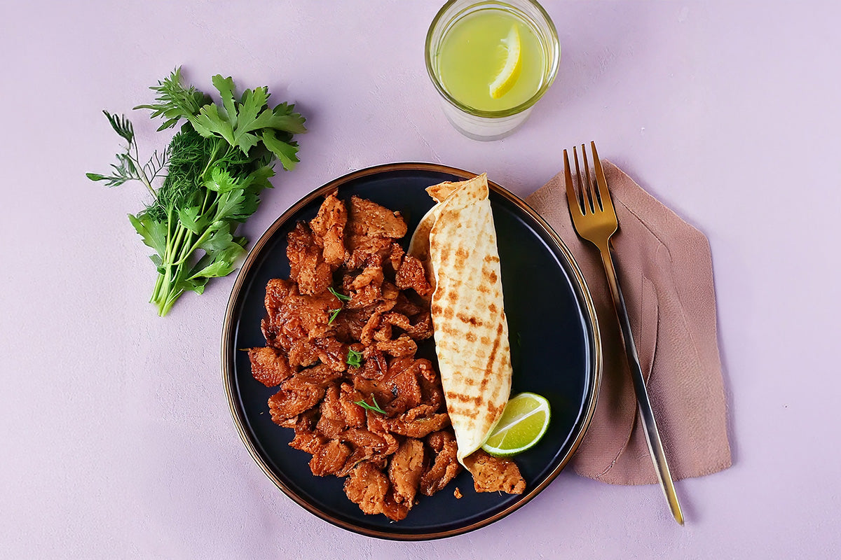 Veganes Gyros aus Sojaprotein auf schwarzem Teller mit Fladenbrot, Limettenscheibe, frischen Kräutern und Glas mit Zitronenwasser