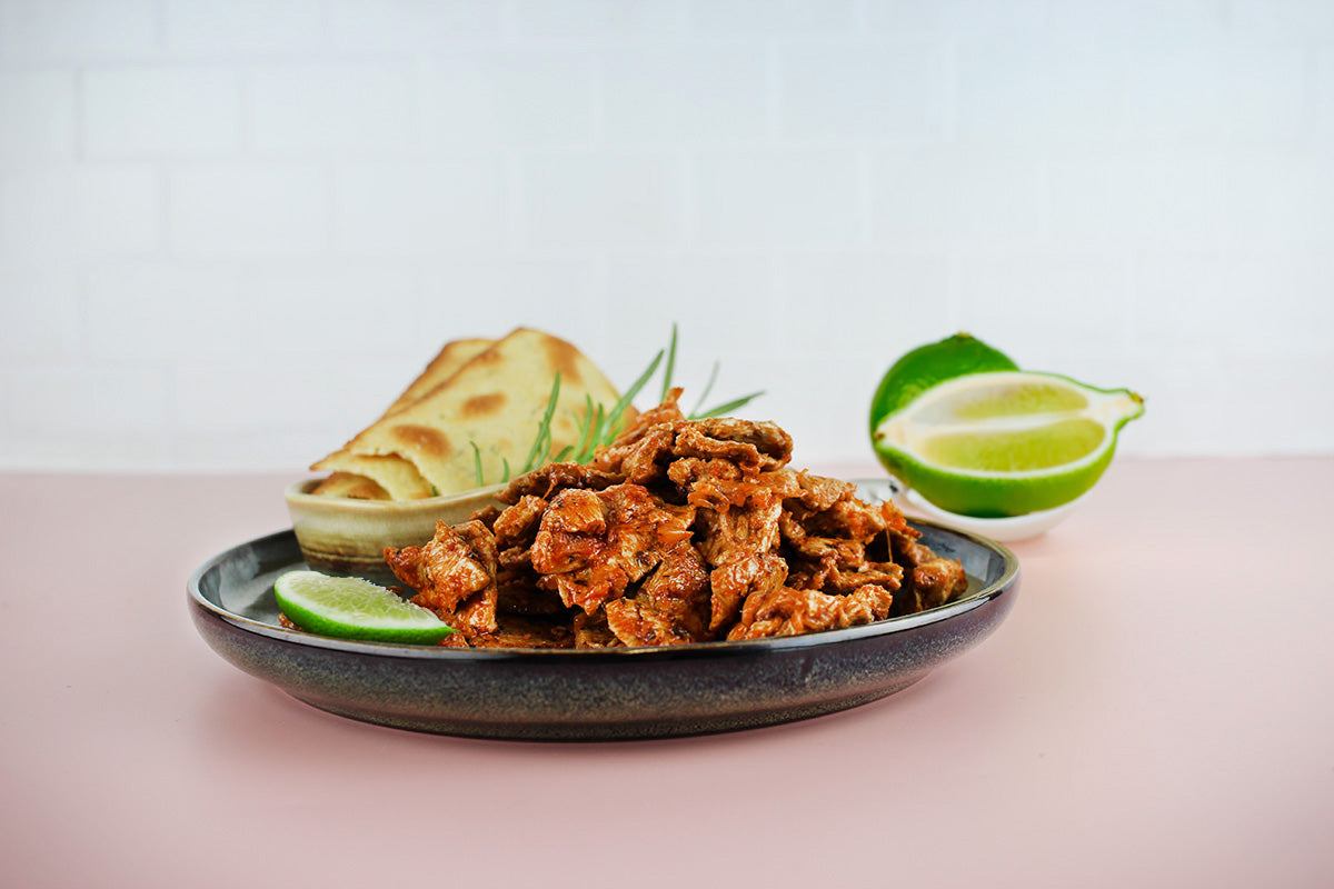 Veganes Gyros aus Sojaprotein auf einem schwarzen Teller mit Limettenscheibe, Fladenbrot und Limettenhälfte im Hintergrund