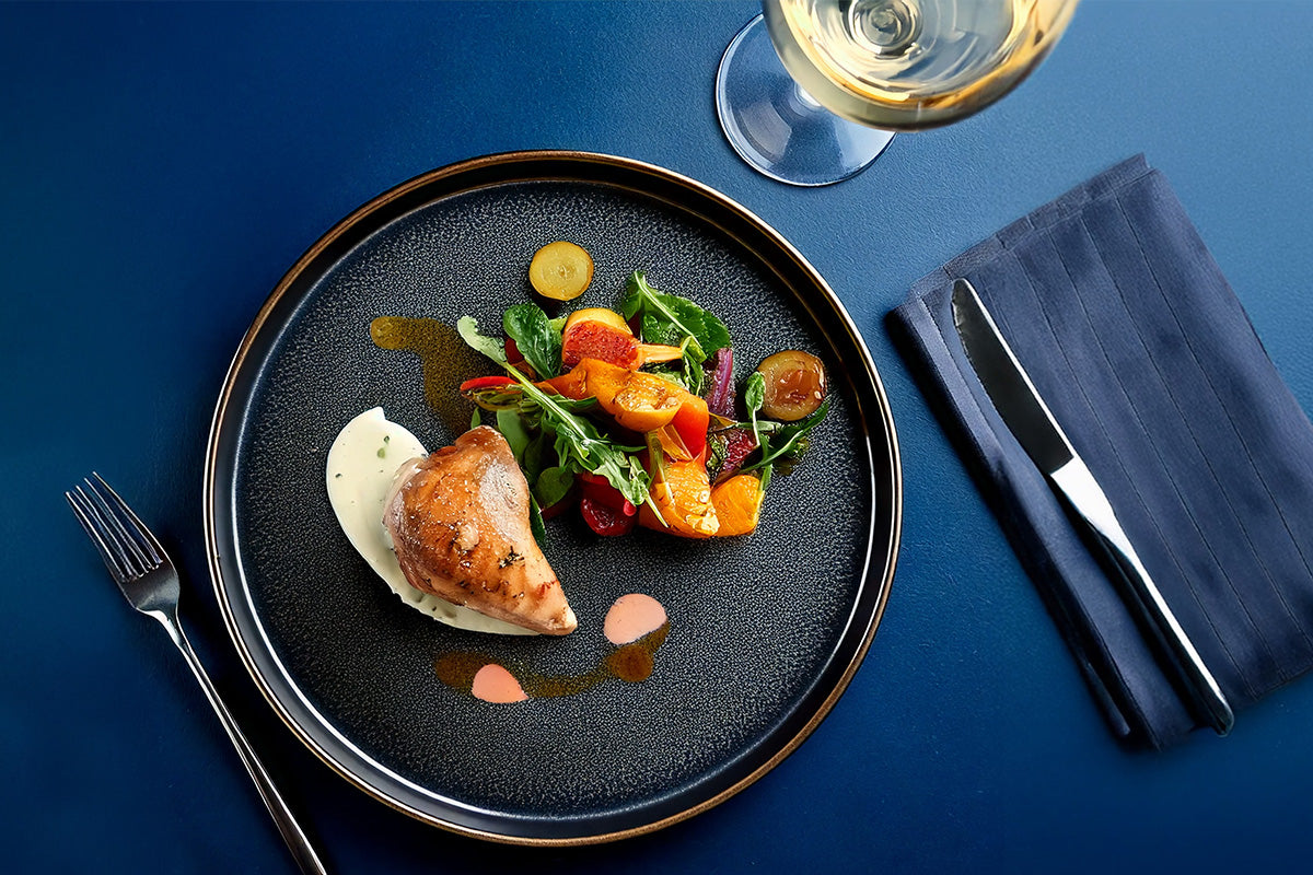 Gegrilltes Wachtelbrustfilet auf schwarzem Teller mit gemischtem Gemüse und zwei Saucen, daneben Besteck auf dunkelblauem Untergrund und ein Glas Weißwein