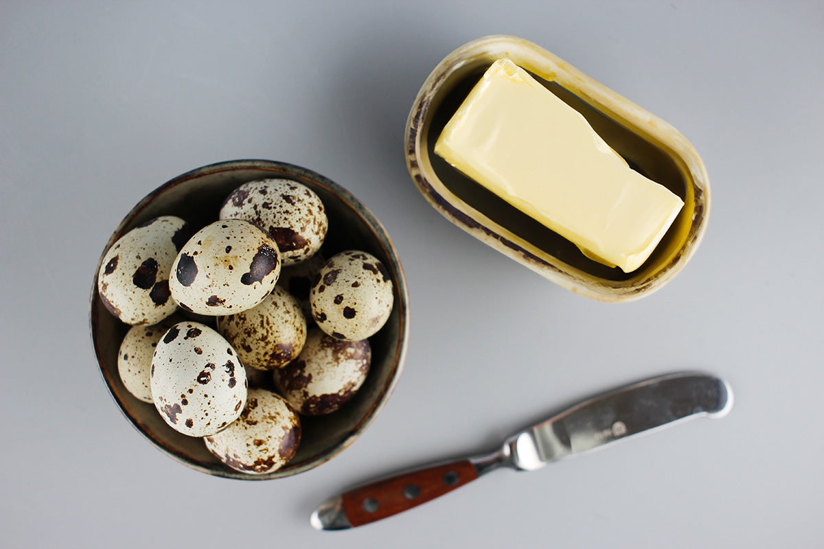 Schale mit gesprenkelten Wachteleiern, Butterstück in Butterdose und Buttermesser auf grauem Untergrund