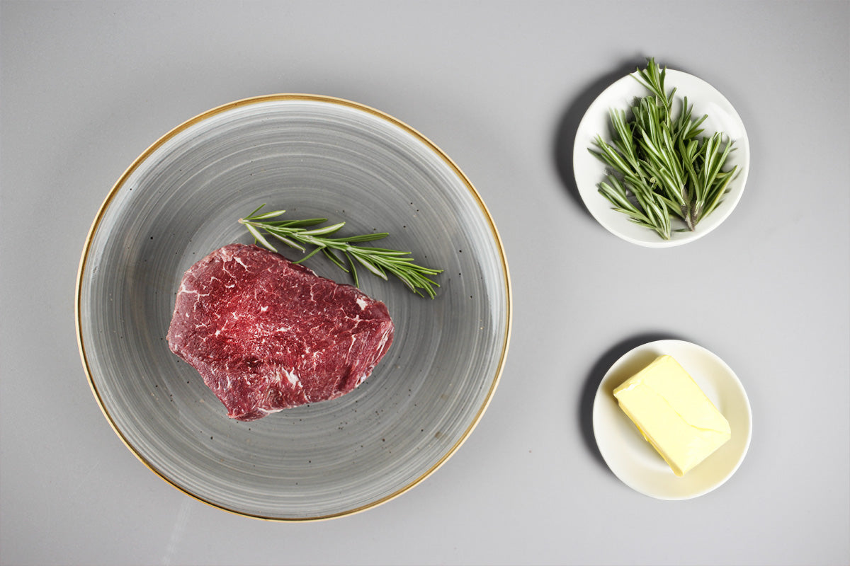 Rohes Wagyu-Rinderfilet auf grauem Teller mit Rosmarinzweig, daneben zwei kleine weiße Teller mit frischem Rosmarin und einem Stück Butter.