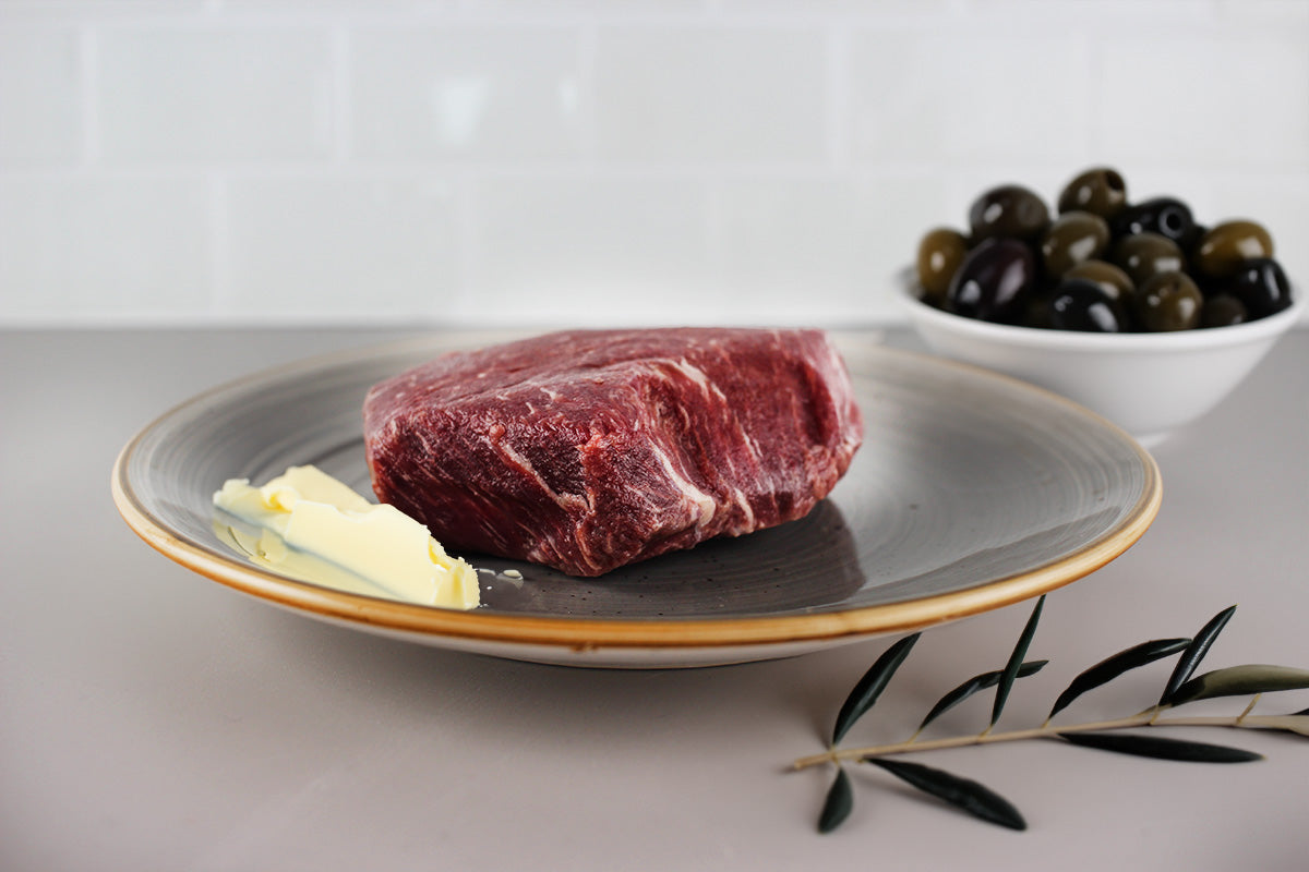 Rohes, marmoriertes Wagyu-Rindfleischstück auf einem hellen Teller mit Basilikumblättern, im Hintergrund eine Schale mit schwarzen Oliven