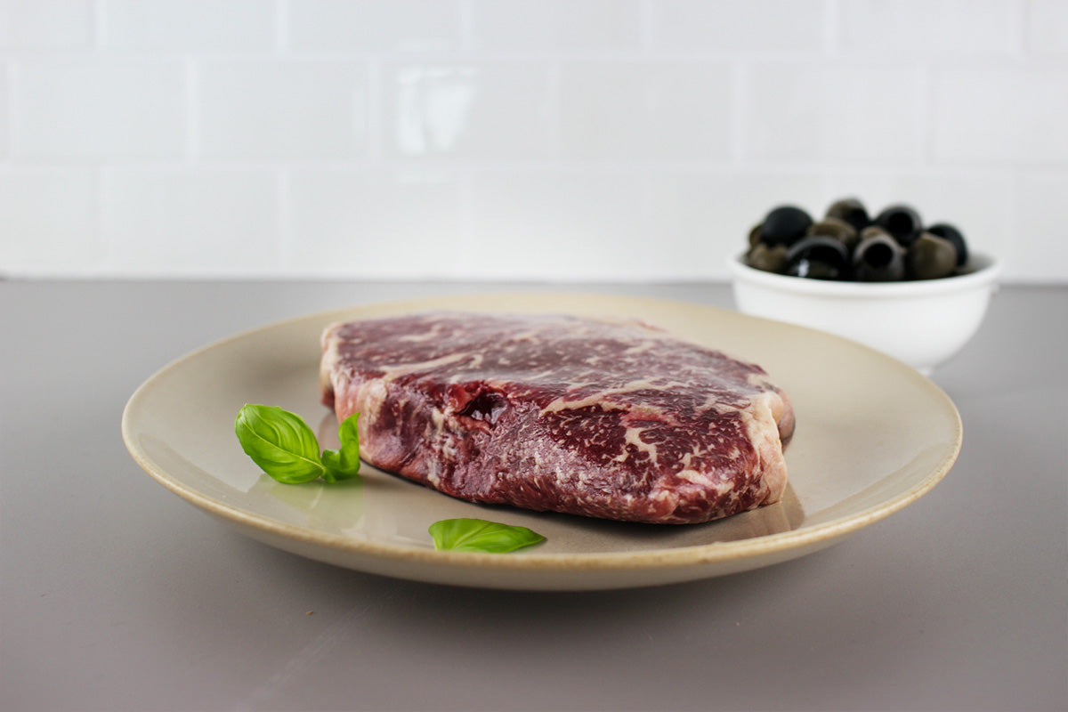 Rohes, marmoriertes Wagyu-Rindfleischstück auf einem hellen Teller mit Basilikumblättern, im Hintergrund eine Schale mit schwarzen Oliven