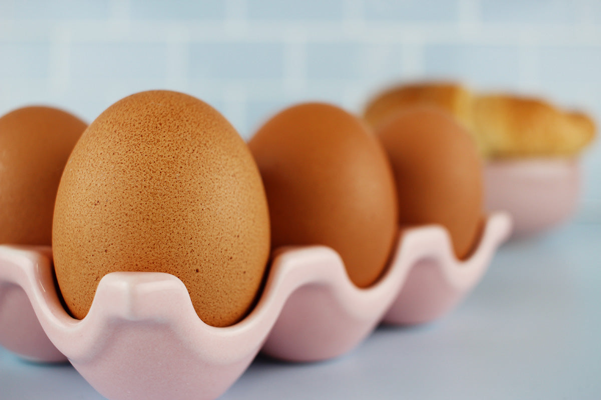 Vier braune Eier in einem rosa Eierhalter, unscharf im Hintergrund ein Croissant auf einem Teller