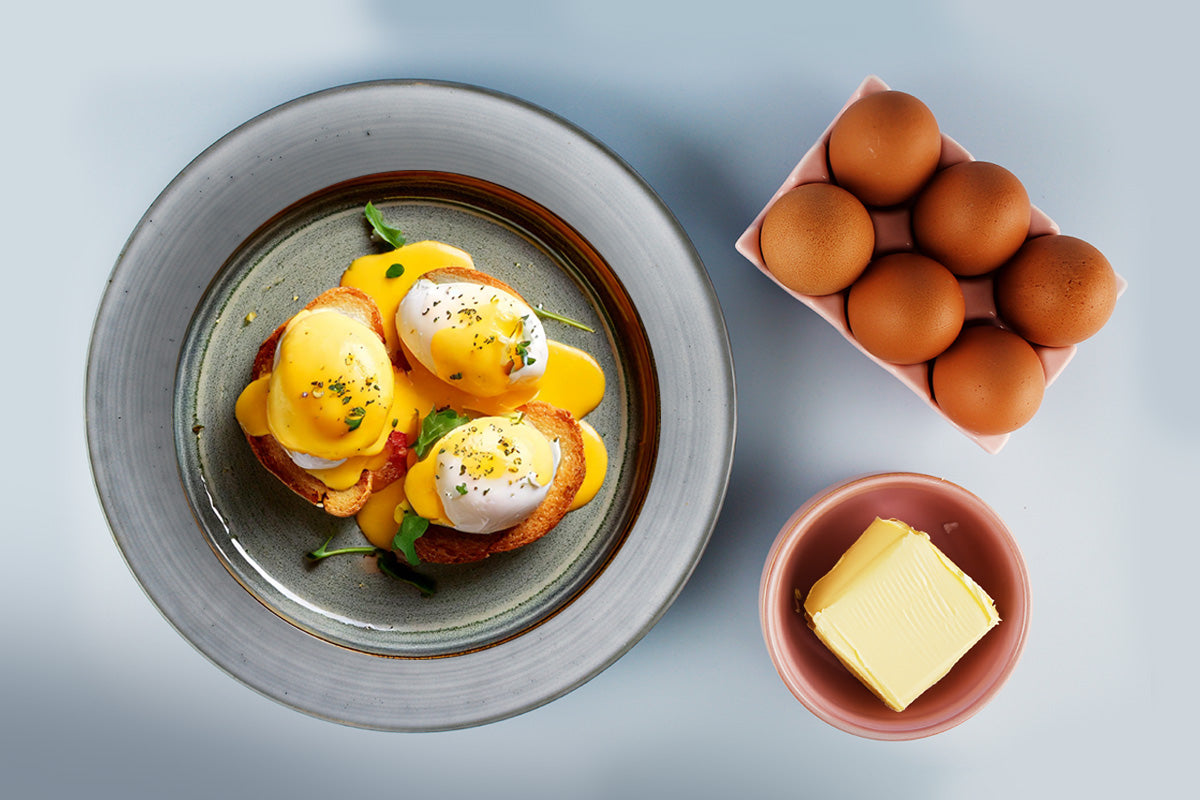 Drei Eggs Benedict mit Sauce Hollandaise auf einem grauen Teller, daneben sechs braune Eier in einem Karton und ein Stück Butter in einer kleinen Schale