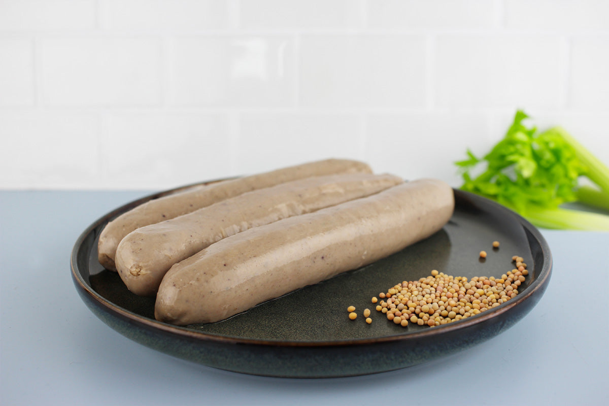 Drei rohe Bratwürste auf einem schwarzen Teller mit Senfkörnern und Sellerie im Hintergrund