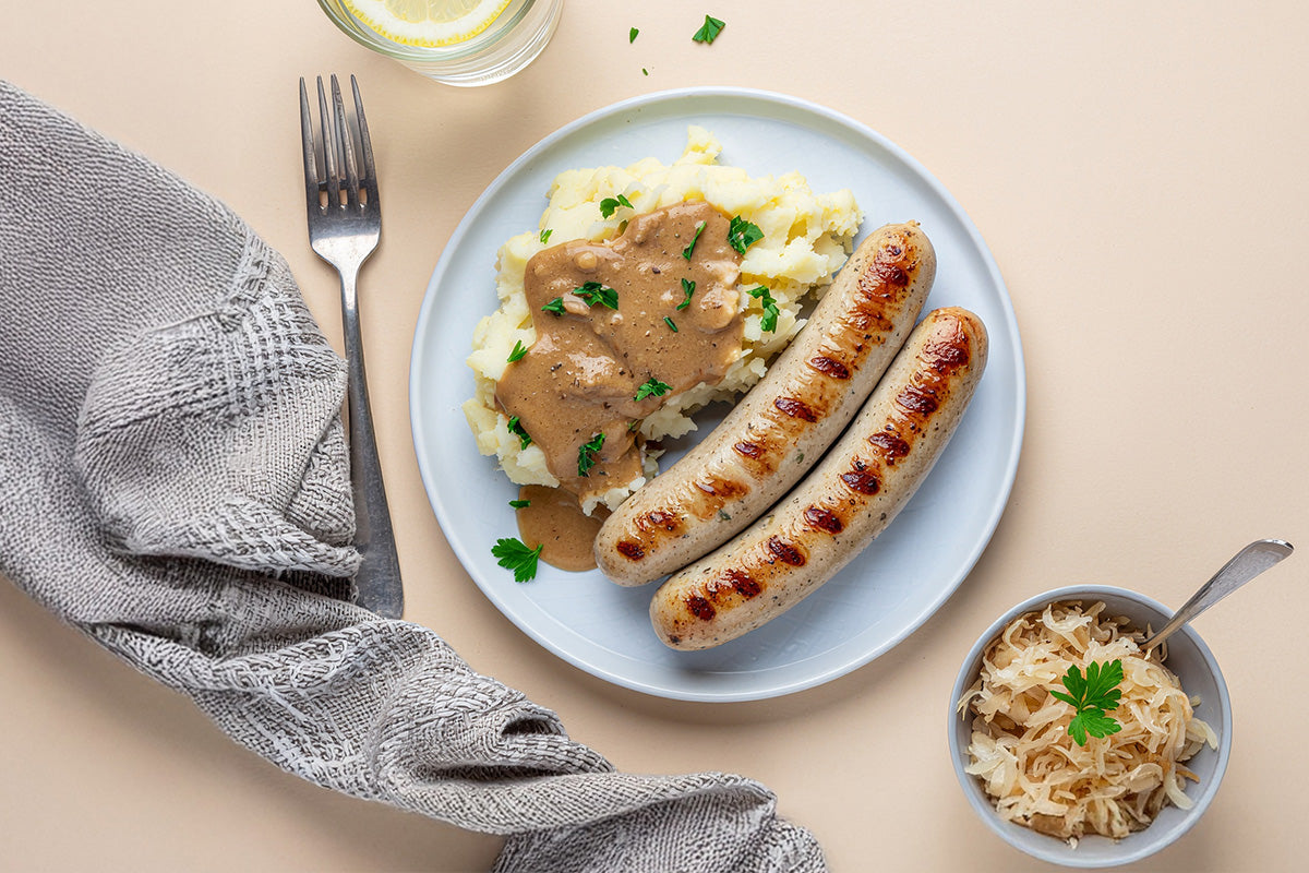 Zwei gegrillte Bratwürste auf einem Teller mit Kartoffelbrei und brauner Soße daneben eine Schale mit Sauerkraut und ein Glas Wasser mit Zitronenscheibe