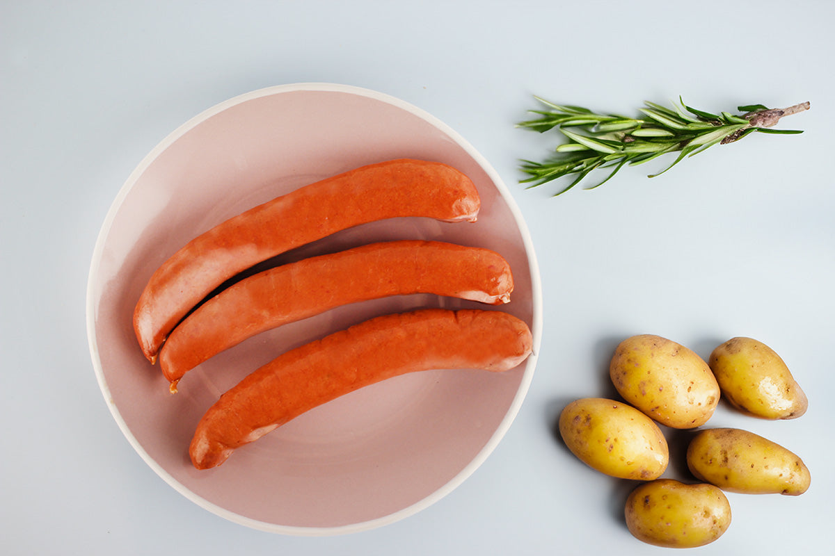 Drei gebogene Würste auf einem runden Teller neben kleinen Kartoffeln und einem Rosmarinzweig auf hellem Untergrund