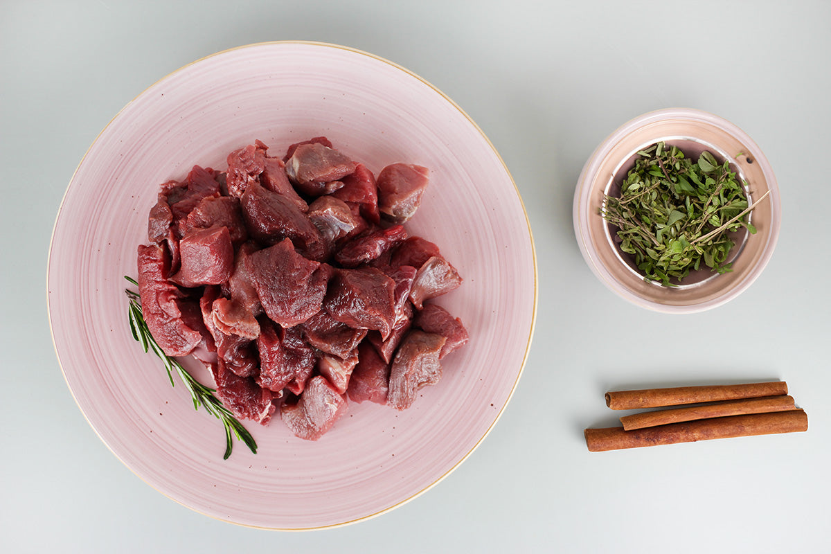 Rohes Wildfleisch in Würfeln auf einem rosa Teller mit Rosmarinzweig, daneben frische Kräuter in einer kleinen Schale und drei Zimtstangen auf weißem Untergrund