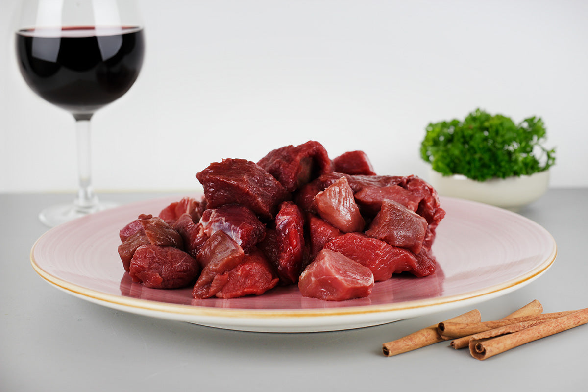 Rohes Wildgulasch auf einem Teller, daneben Zimtstangen, ein Glas Rotwein und eine Schale mit Petersilie