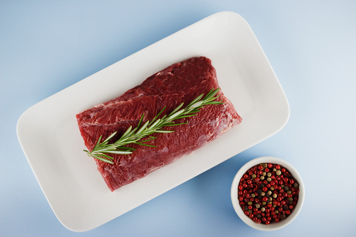 Roher Wildschweinrücken auf weißem Teller mit Rosmarinzweig, daneben Schale mit bunten Pfefferkörnern auf blauem Hintergrund