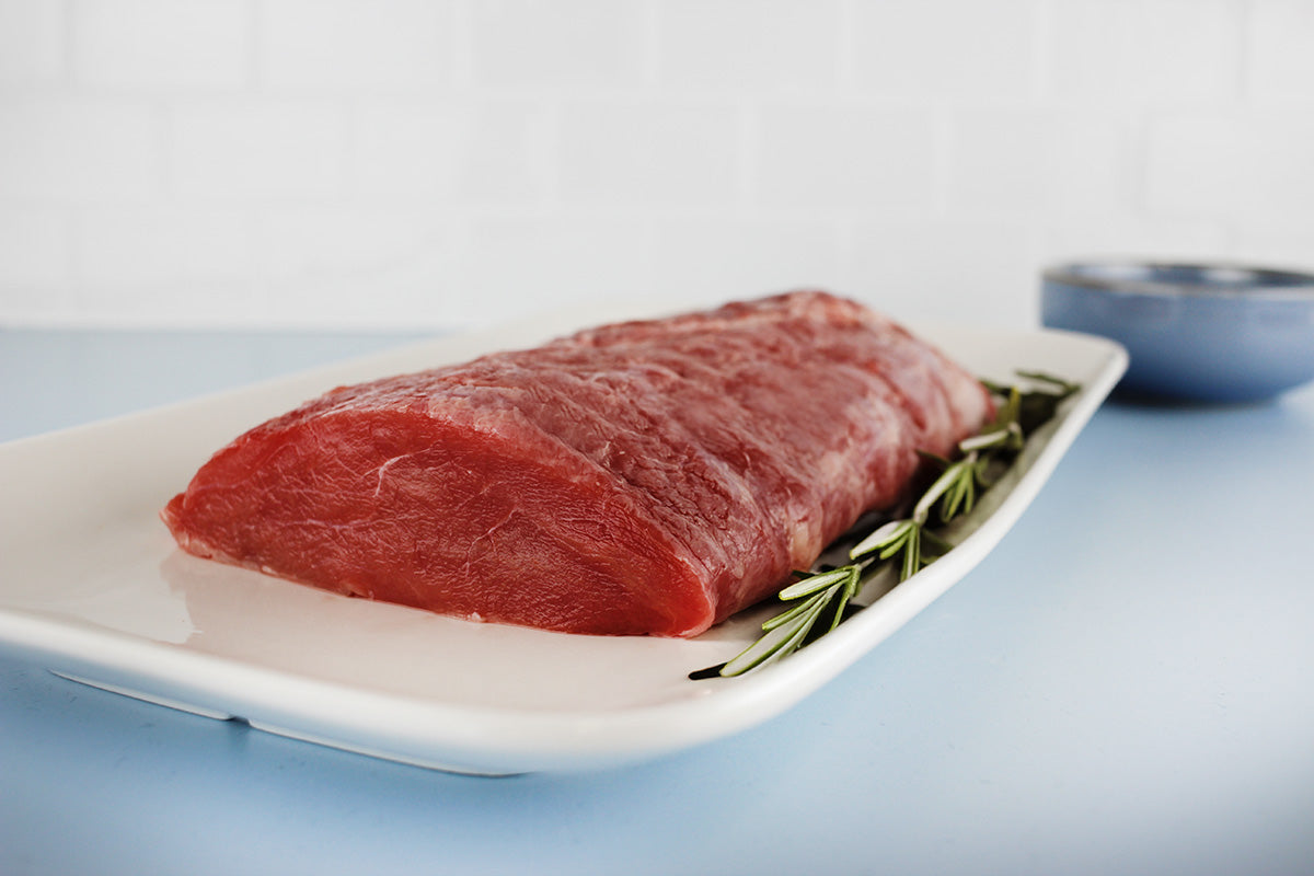 Roher Wildschweinrücken auf weißem Teller mit Rosmarinzweigen, vor hellem Hintergrund