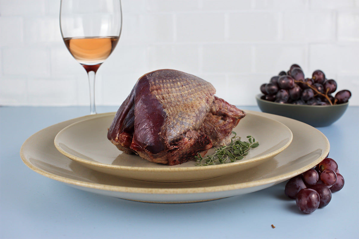 Wildtaube auf einem Teller mit frischen Kräutern, daneben eine Schale und Trauben, im Hintergrund ein Glas Roséwein