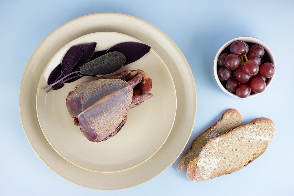 Ausgenommene rohe Wildtaube mit Salbeiblättern auf einem Teller, daneben zwei Scheiben Brot und eine Schale mit roten Trauben auf blauem Untergrund