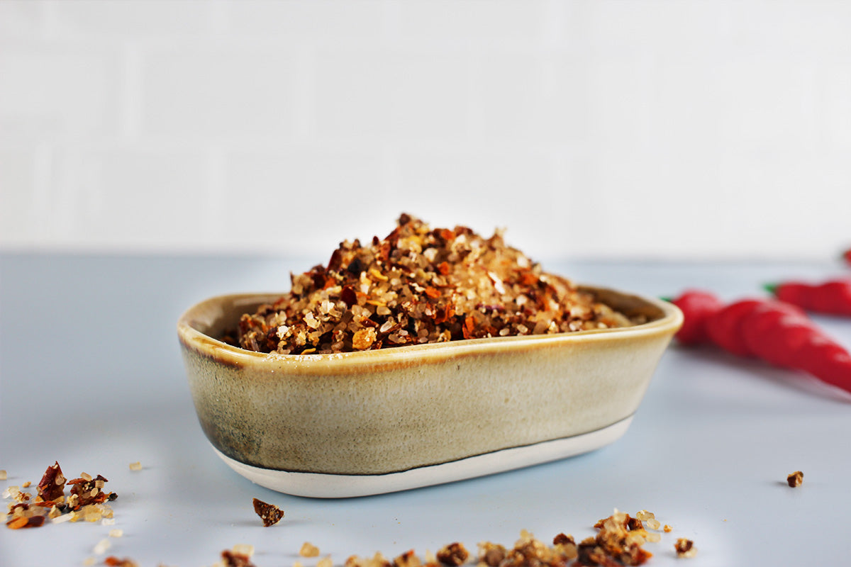 Braune Keramikschale mit grob zerstoßenen Chiliflocken, verstreut auf weißer Oberfläche, rotes Tuch im Hintergrund.