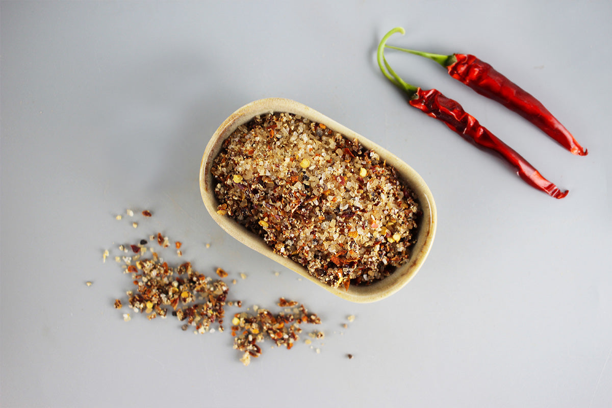 Kleines Holzschälchen mit grobem Gewürzsalz und zwei roten getrockneten Chilischoten mit grünen Stielen auf grauem Untergrund.