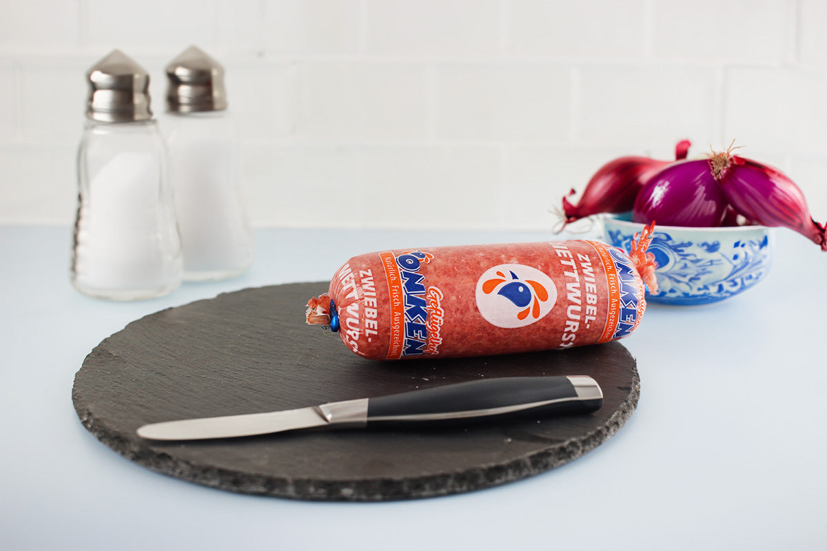 Zwiebelmettwurst in Verpackung auf rundem schwarzen Schieferbrett mit Messer, Salzstreuer und rohen roten Zwiebeln in blau-weißem Keramikschälchen