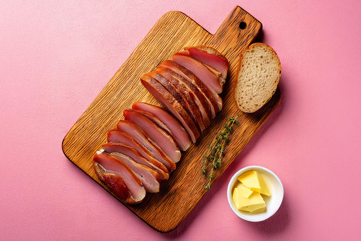 Aufgeschnittene geräucherte Entenbrust auf einem Holzbrett mit einem Stück Brot, Butter in einer kleinen Schale und einem Zweig Thymian auf rosa Untergrund