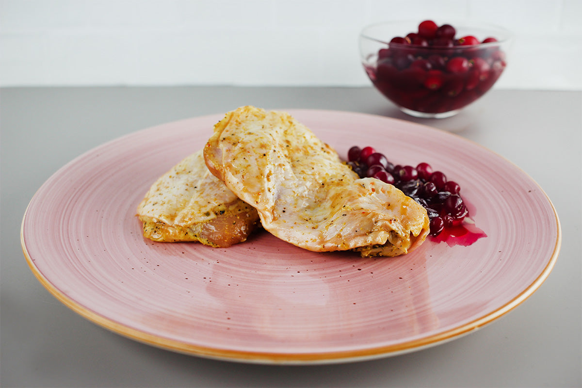 Zwei gebratene Hähnchensteaks auf rosa Teller mit Preiselbeersoße, im Hintergrund Schale mit frischen Preiselbeeren