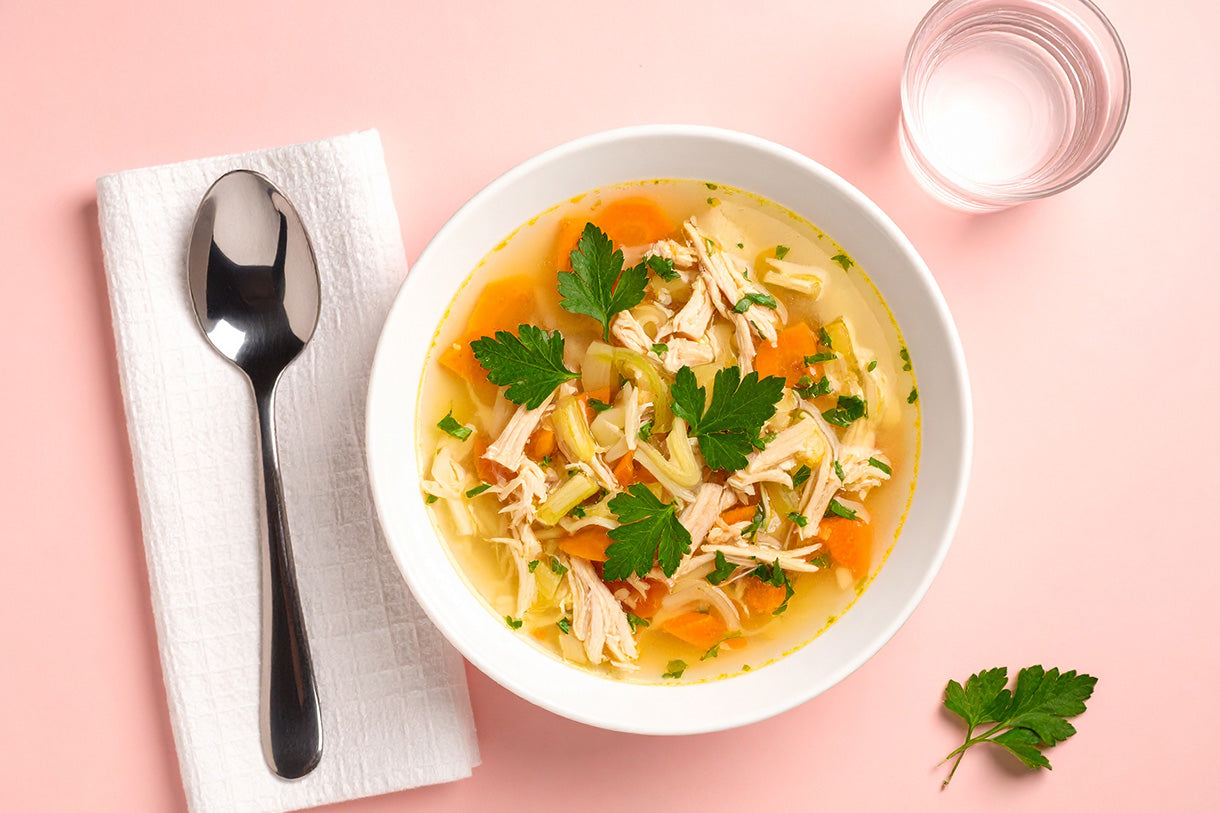 Weiße Schüssel mit Hühnersuppe, Karotten, Sellerie, Hühnerfleisch und Petersilie, daneben Löffel auf weißem Tuch und Glas Wasser auf rosa Untergrund