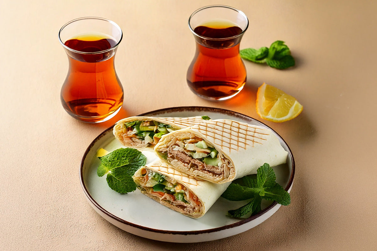 Ein Teller mit drei Wraps gefüllt mit Gemüse und Fleisch, garniert mit Minze, dazu zwei Gläser Tee und eine Zitronenscheibe.